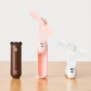 Three JISULIFE portable bear fans in brown, pink, and white, displayed on a wooden table. The brown fan is closed, the pink one is open as a handheld fan, and the white one is folded into a desktop fan position.